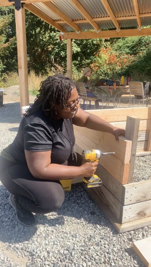 Evans squatting as she uses a drill to attach wooden boards to a post.