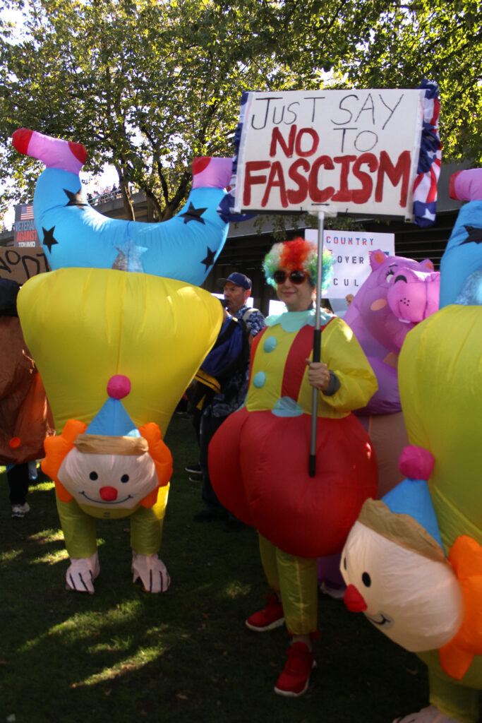 In front of the Morrison Bridge, two matching clowns flank another protestor in a different clown costume holding an anti-fascist sign.