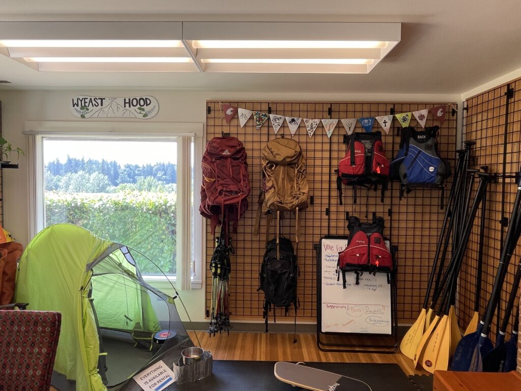 An indoor photo showing a wall of various gear, including life jackets, paddle boards and more, as well as a tent to the left of the other gear. Photo is taken by Ashlynn Fredendall.