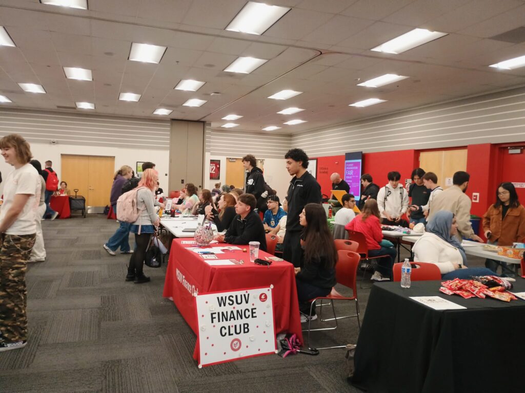 Image contains a crowded room, the photo is focused on a table with a red covering, and on its side a poster with the words "WSUV Finance Club."